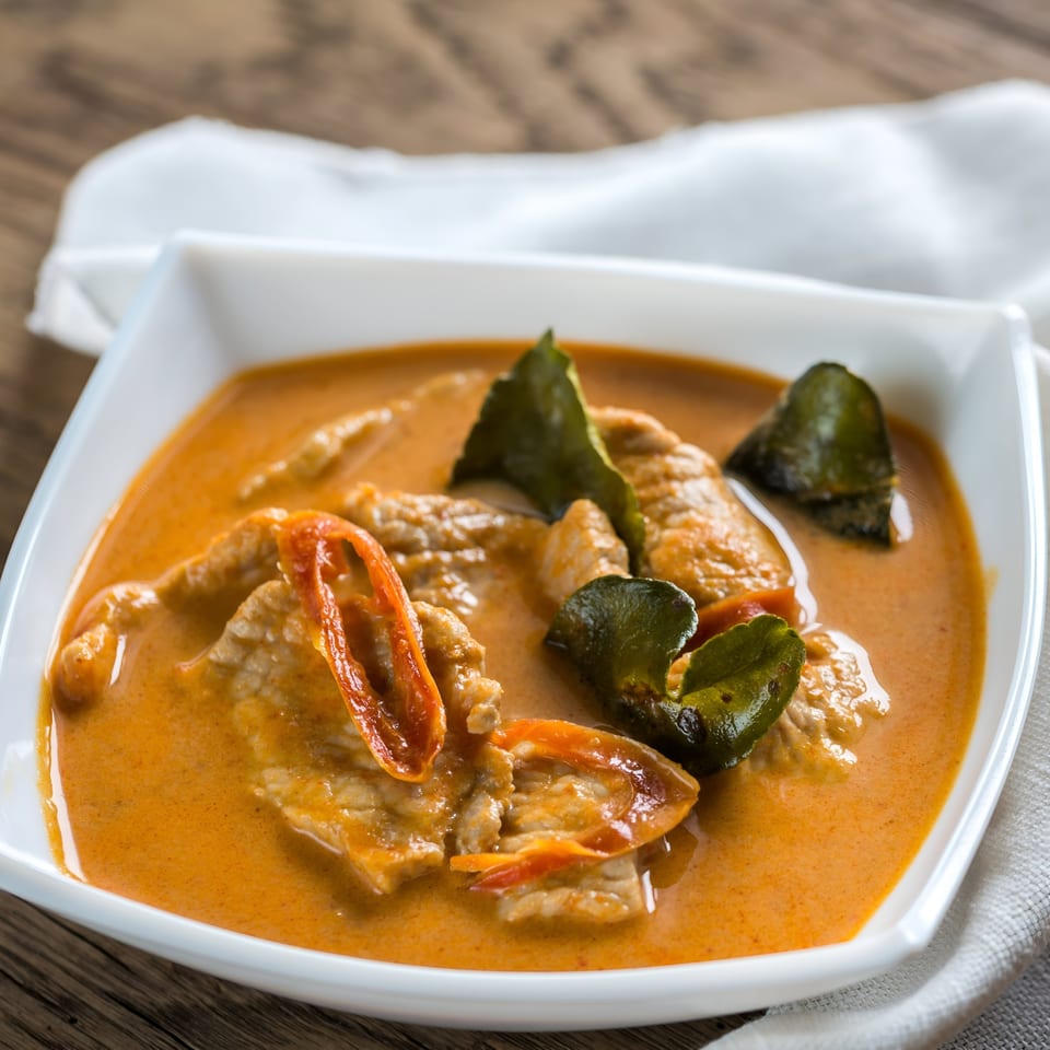 Close-up of Kati Thai Eatery's red curry with meat, chili, and kaffir lime leaves in a white bowl.